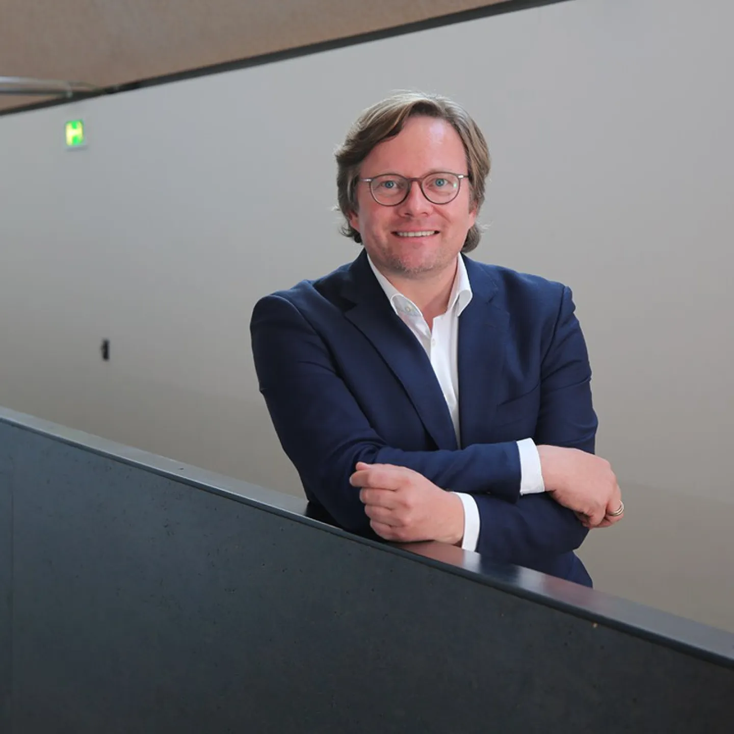 Ein Mann in einem blauen Anzug lehnt lächelnd an einem Geländer in einem modernen Gebäude.