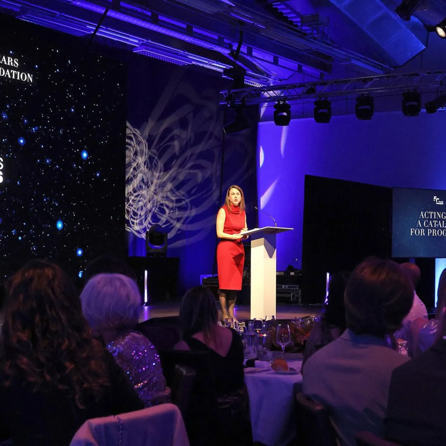 Eine Rednerin in rotem Kleid steht an einem Podium auf einer Bühne, umgeben von Publikum. Im Hintergrund ist eine Leinwand mit der Aufschrift "Celebrating 50 Years of the Onassis Foundation" zu sehen. Die Veranstaltung findet in einem festlich beleuchteten Raum statt.