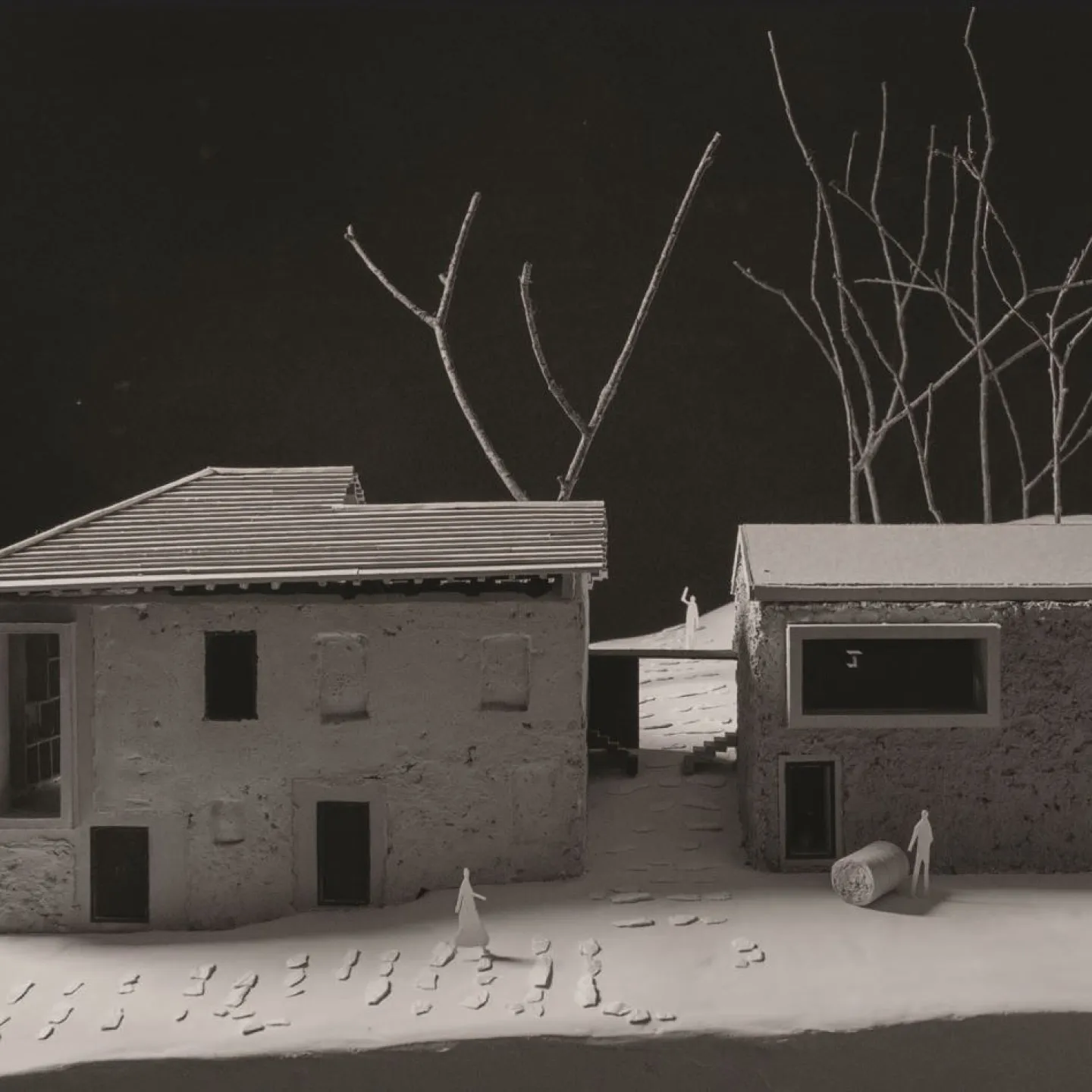 Modell eines architektonischen Projekts mit zwei Gebäuden, umgeben von kahlen Bäumen. Das linke Gebäude hat ein geneigtes Dach, während das rechte ein Flachdach aufweist. Kleine Figuren und Fußspuren sind auf dem Boden sichtbar, was eine menschliche Präsenz andeutet.