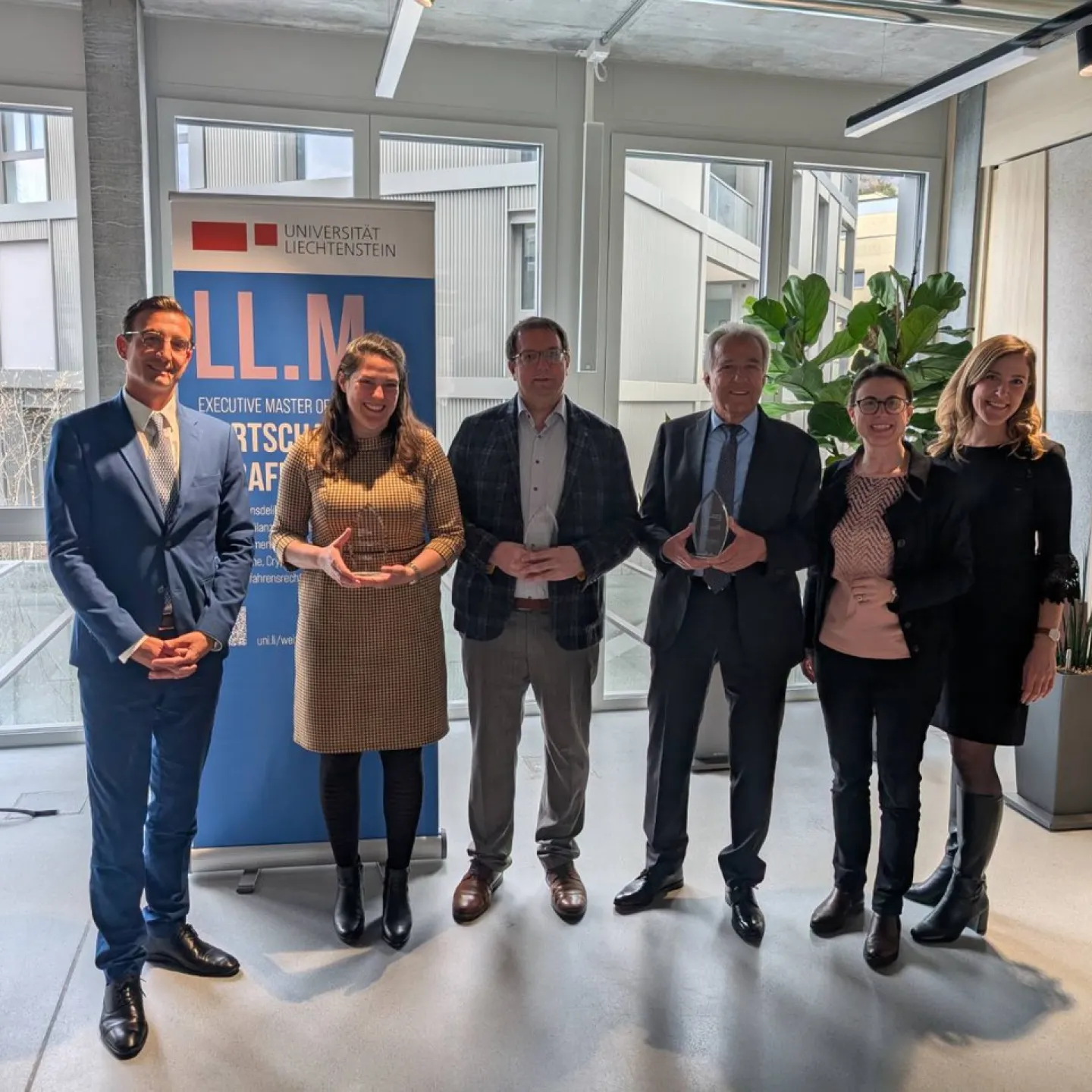 Sechs Personen stehen in einem modernen Raum vor einem Banner der Universität Liechtenstein. Zwei Personen halten Glastrophäen. Die Gruppe lächelt in die Kamera. Im Hintergrund sind große Fenster und Pflanzen zu sehen.