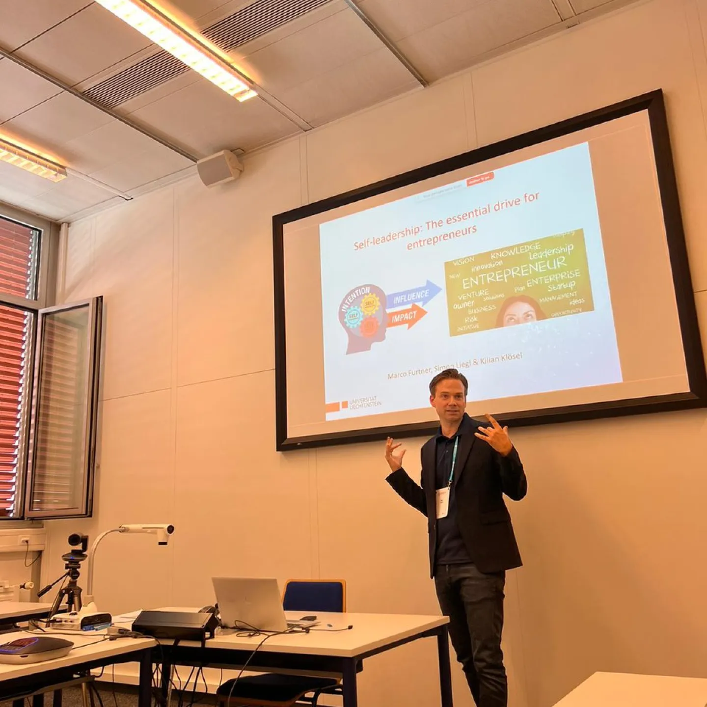 Ein Vortragender präsentiert in einem Seminarraum ein Paper über Selbstführung für Unternehmer. Im Hintergrund ist eine Leinwand mit einer Präsentationsfolie zu sehen. Tische mit Laptops und Kamera sind im Vordergrund.