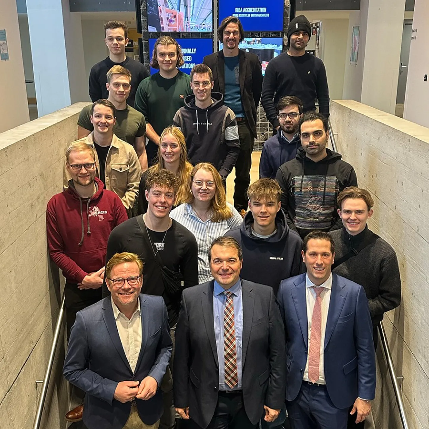 Gruppe von Studierenden und Lehrenden steht auf einer Treppe in einem modernen Gebäude der Universität Liechtenstein, lächelnd in die Kamera. Im Hintergrund sind Bildschirme und eine Steinwand zu sehen.