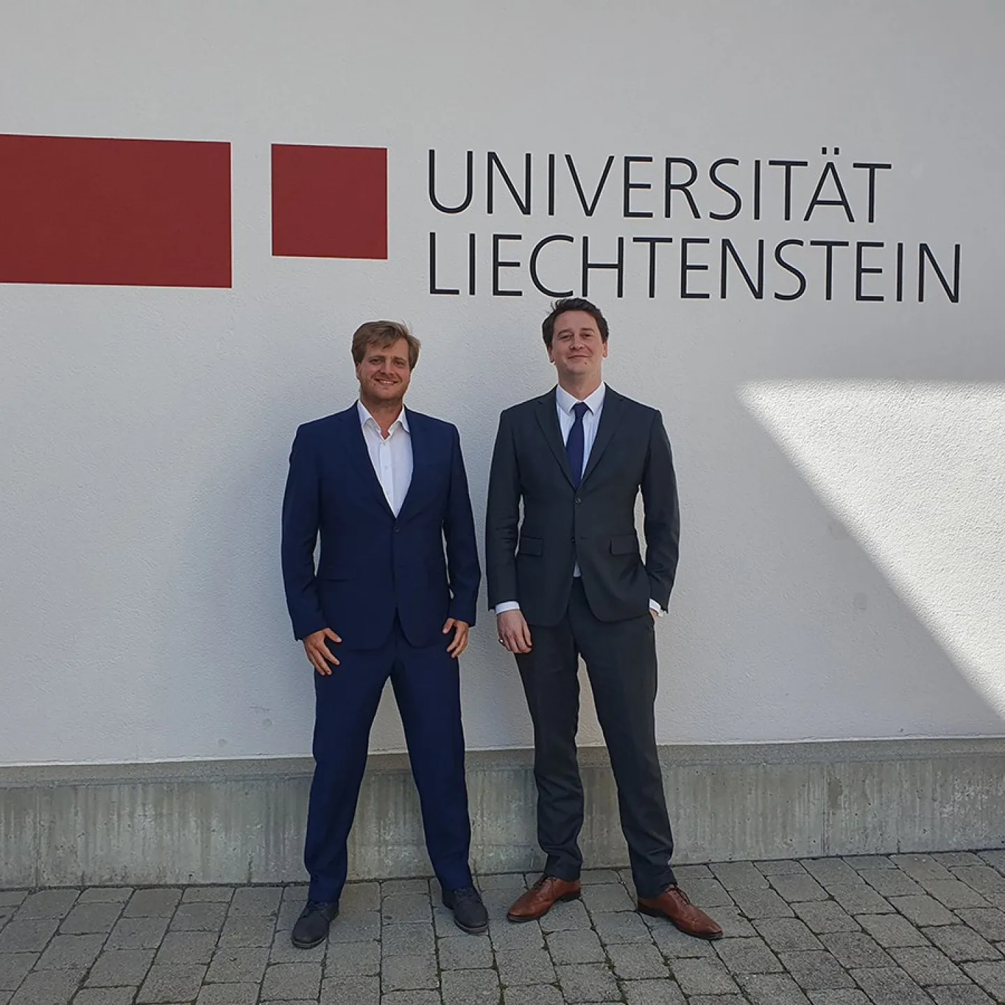 Zwei Personen in Anzügen stehen vor einer Wand mit dem Logo der Universität Liechtenstein.