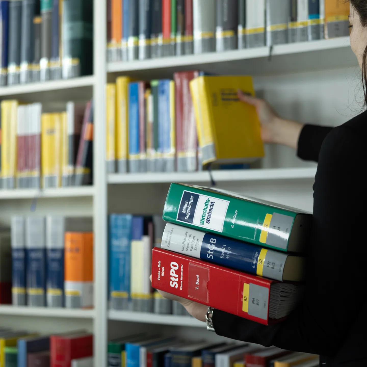 Frau stellt gelbes Fachbuch ins Regal und hält weitere juristische Bücher in der Bibliothek der Universität Liechtenstein.