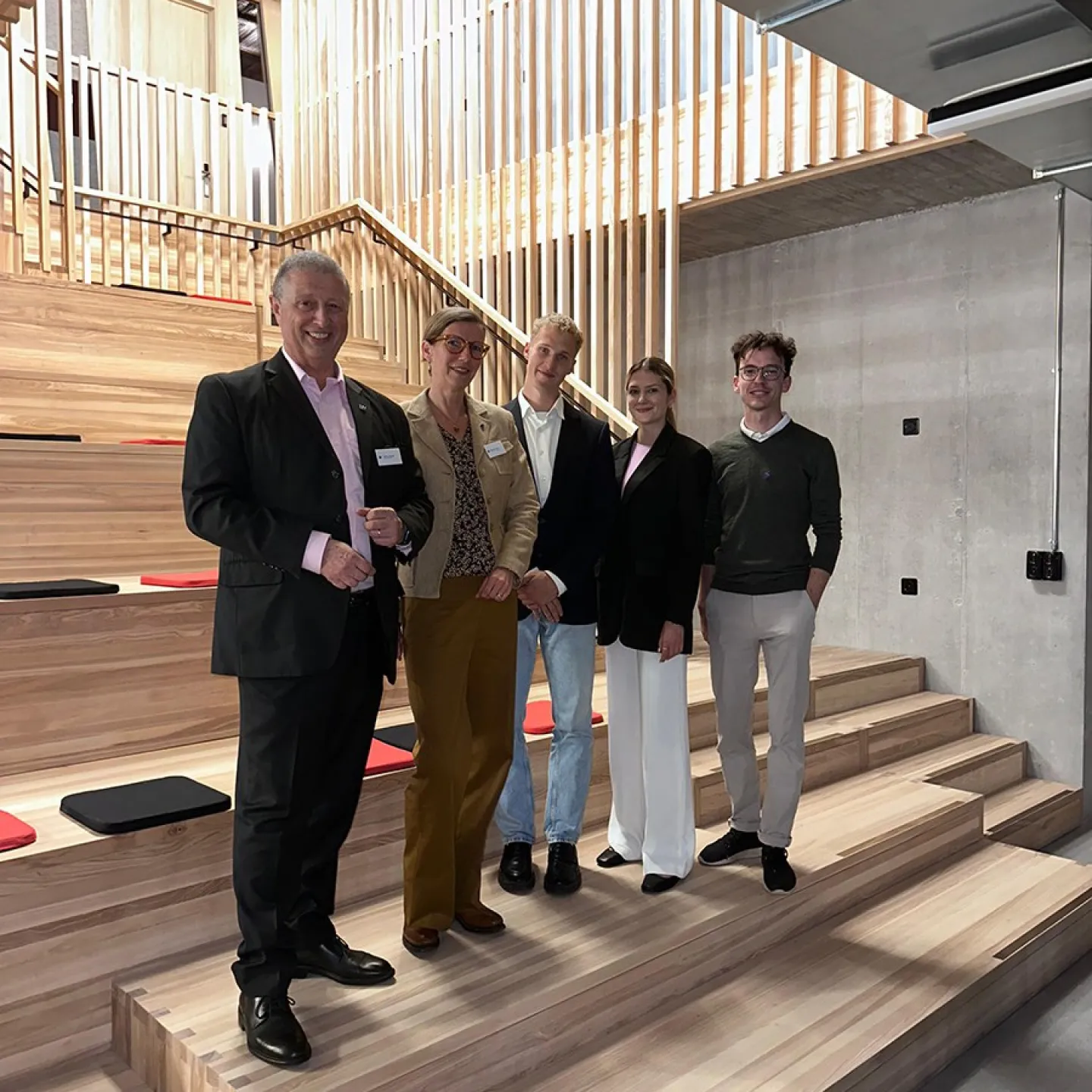 Fünf Personen stehen lächelnd in einer modernen Aula mit Holzstufen und vertikalen Holzlamellen im Hintergrund. Sie tragen formelle Kleidung und posieren für ein Gruppenfoto.