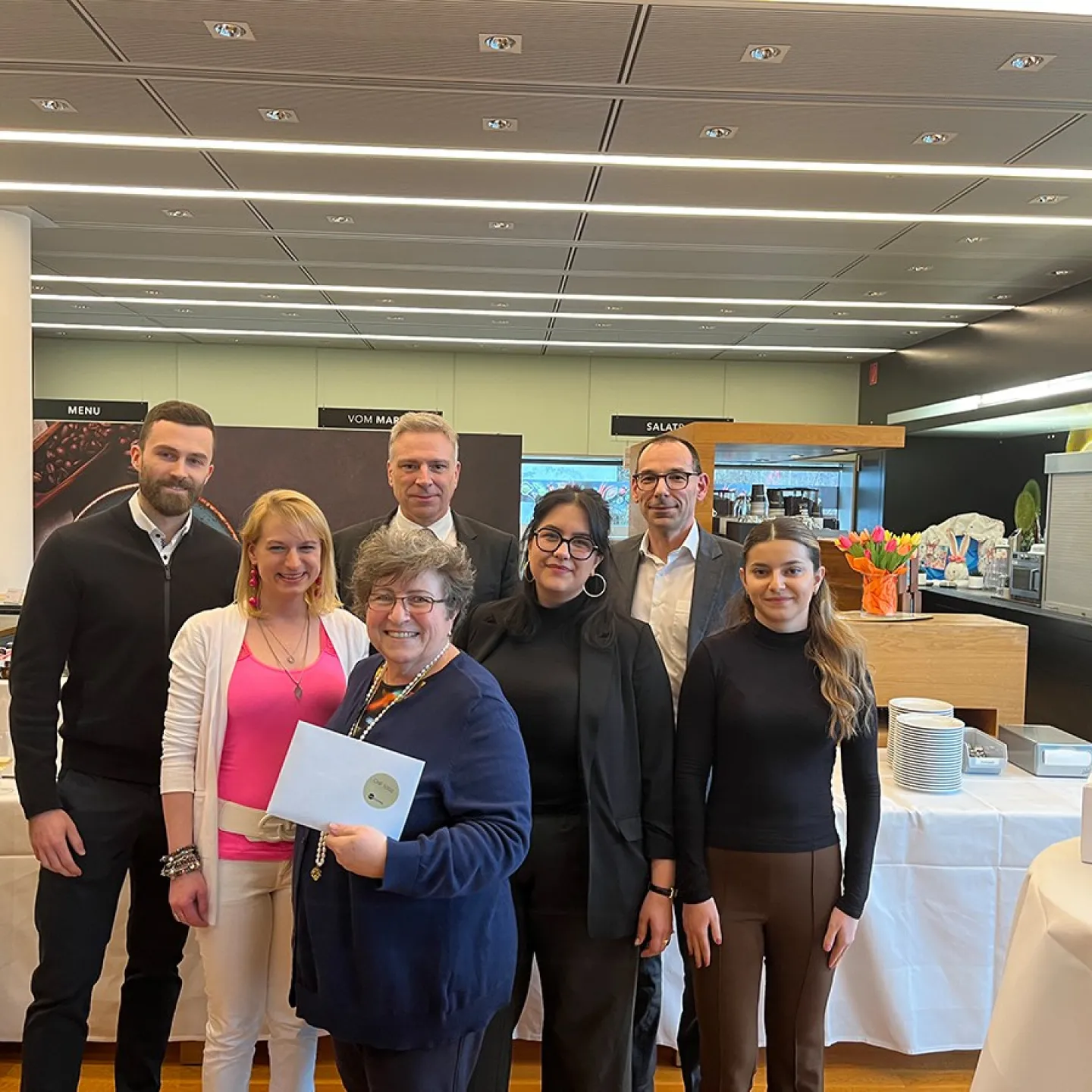 Eine Gruppe von sieben Personen steht lächelnd in einem modernen Raum. Eine Person hält ein Zertifikat in den Händen. Im Hintergrund sind Tische mit Geschirr und Dekorationen zu sehen.