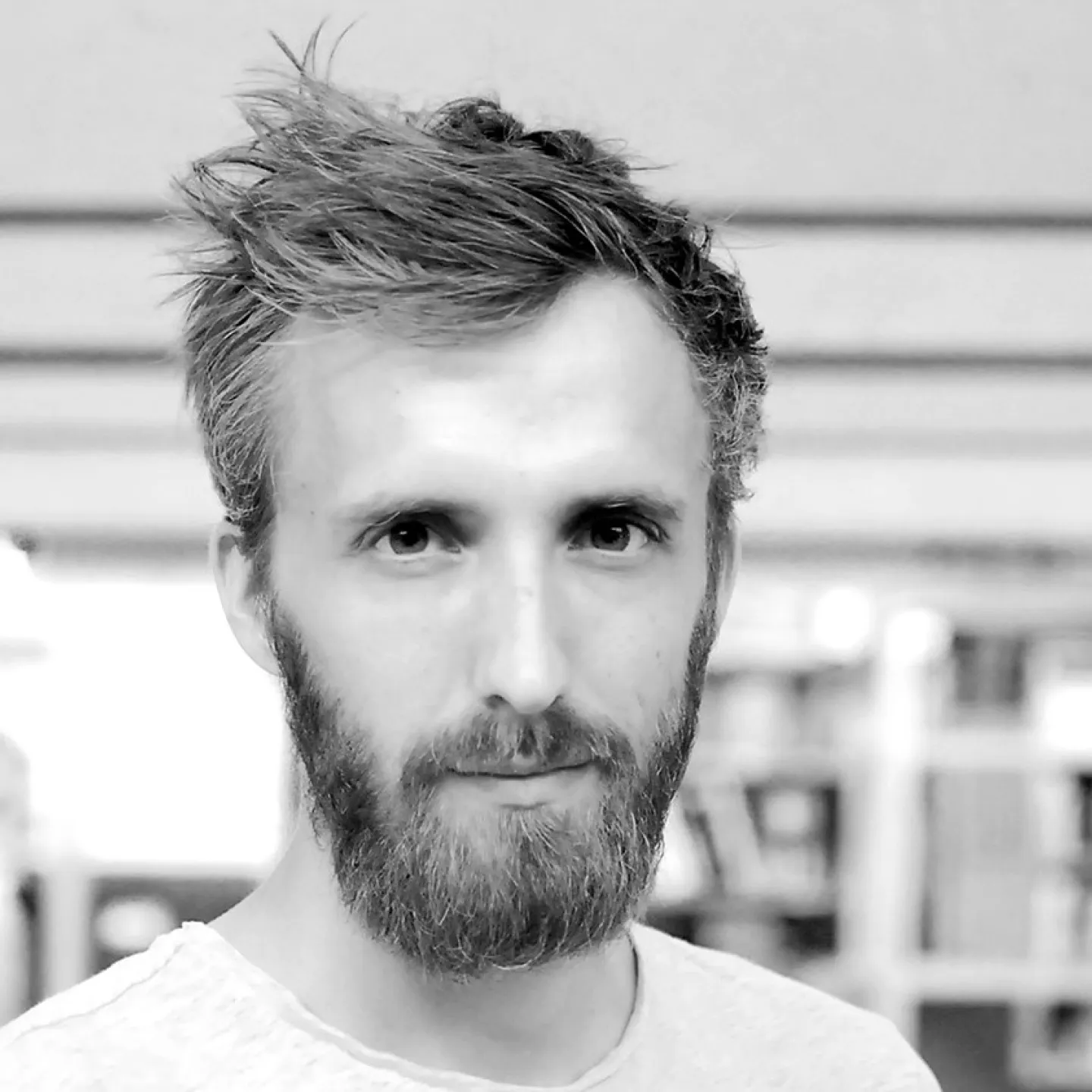 Ein Mann mit Bart und kurzem Haar blickt direkt in die Kamera. Der Hintergrund ist unscharf und zeigt eine Innenraumszene.