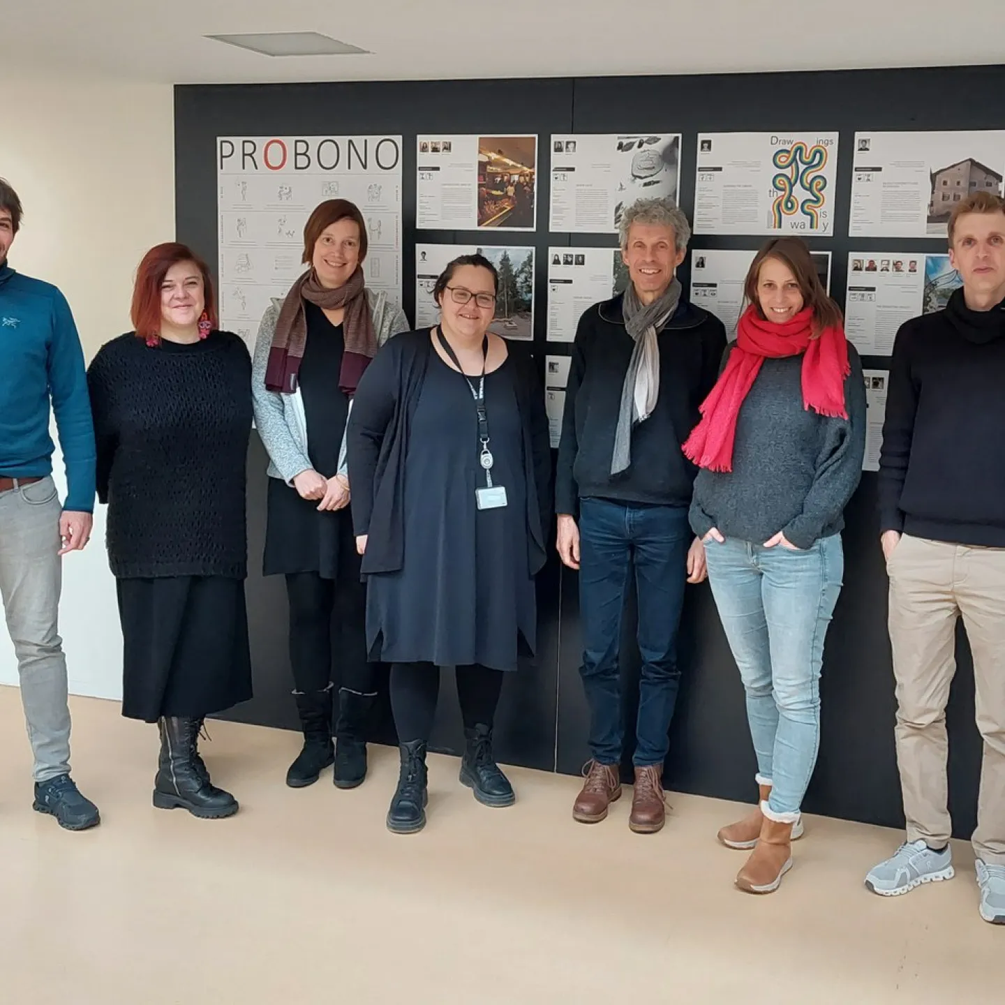 Gruppe von sieben Personen steht vor einer Wand mit dem Titel "PROBONO" und mehreren Plakaten. Sie lächeln in die Kamera und repräsentieren das Service Learning Netzwerk an der Universität Liechtenstein.