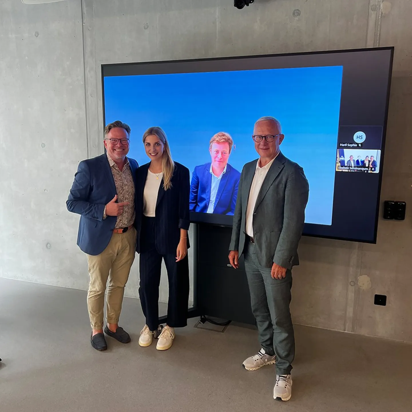 Drei Personen stehen vor einem Bildschirm, der zwei weitere Personen in einem virtuellen Meeting zeigt. Die Szene befindet sich in einem modernen Raum mit Betonwänden.