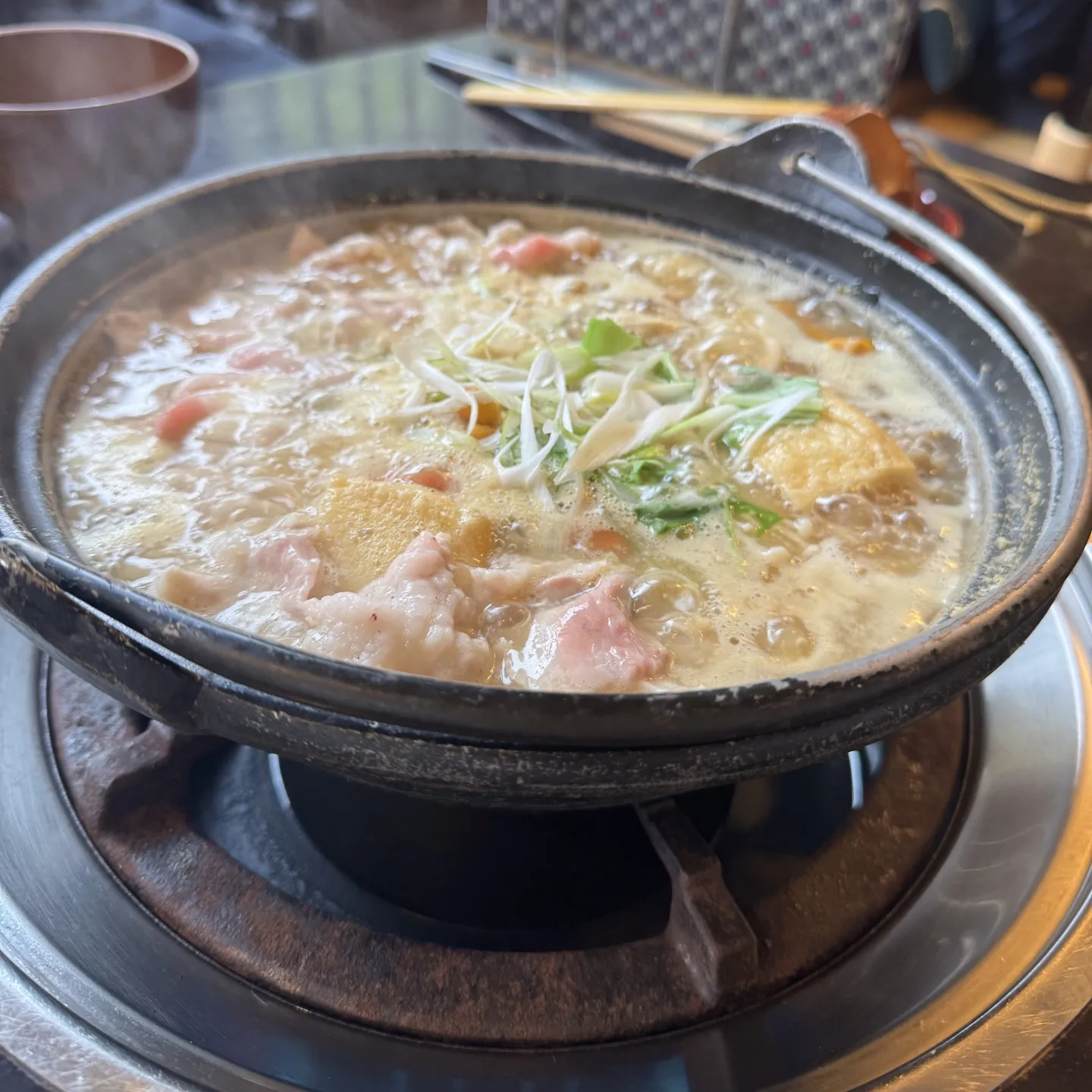 Dampfender Hot Pot mit Fleisch, Tofu und Gemüse in heller Brühe auf einem Tisch-Gasbrenner.