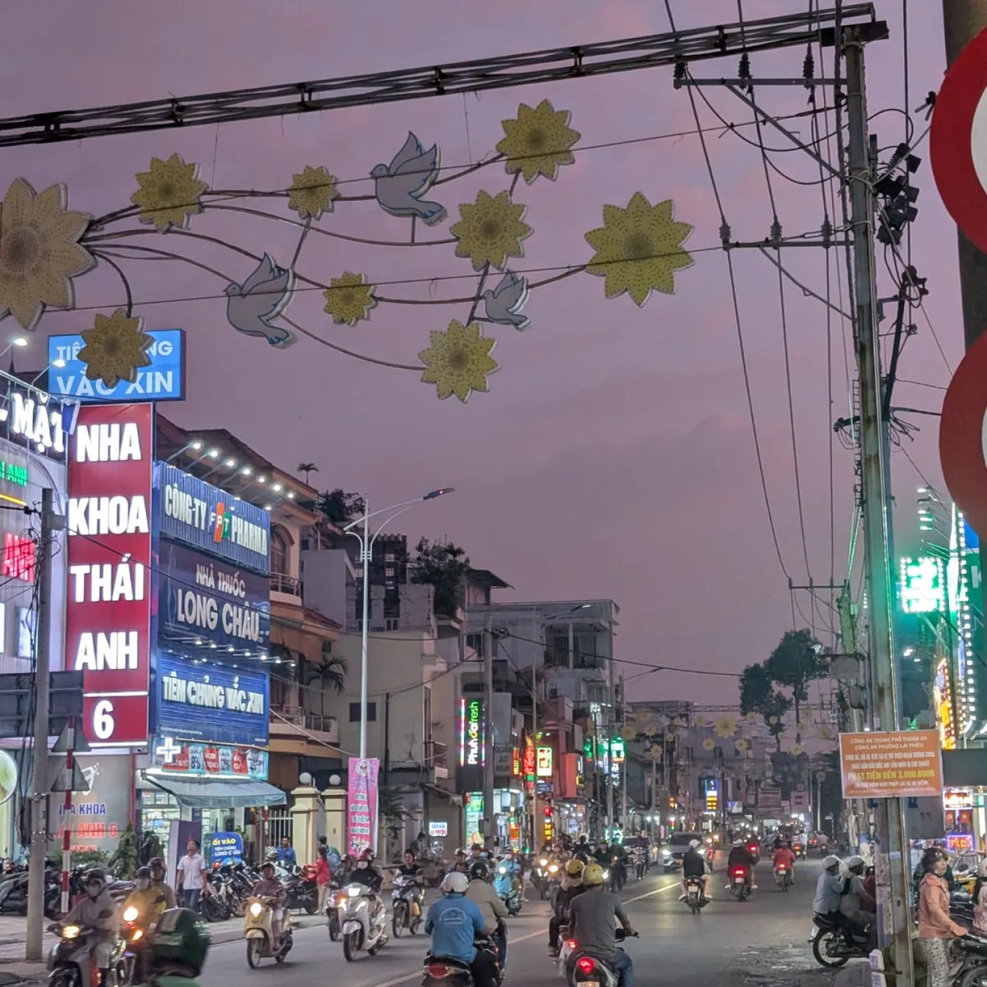 Belebte Straße bei Dämmerung mit vielen Motorrollern, leuchtenden Werbeschildern und dekorativen Lichtern über der Fahrbahn.
