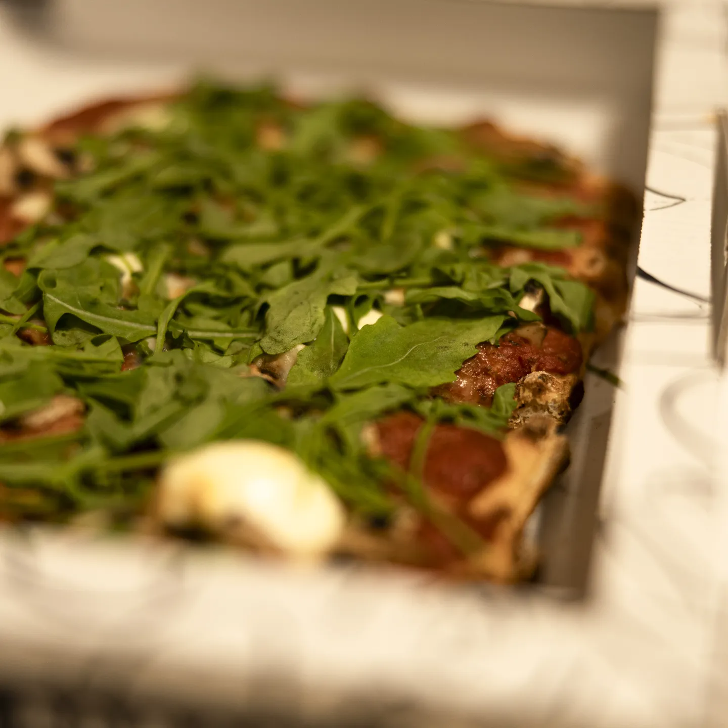 Detailaufnahme einer Pizza mit Rucola und Pilzen in einer geöffneten Pizzaschachtel.