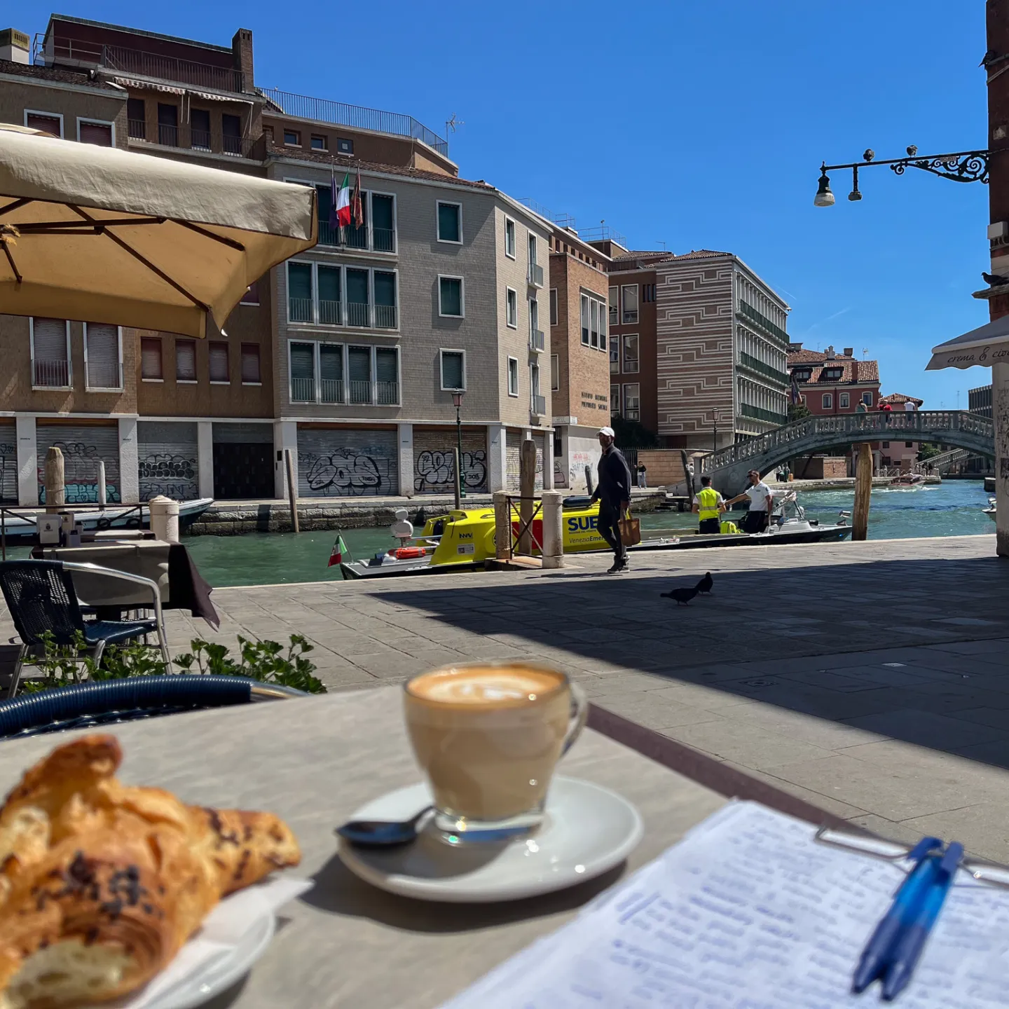 Kaffee, Croissant und Notizen auf einem Tisch an einem Kanal in Venedig.