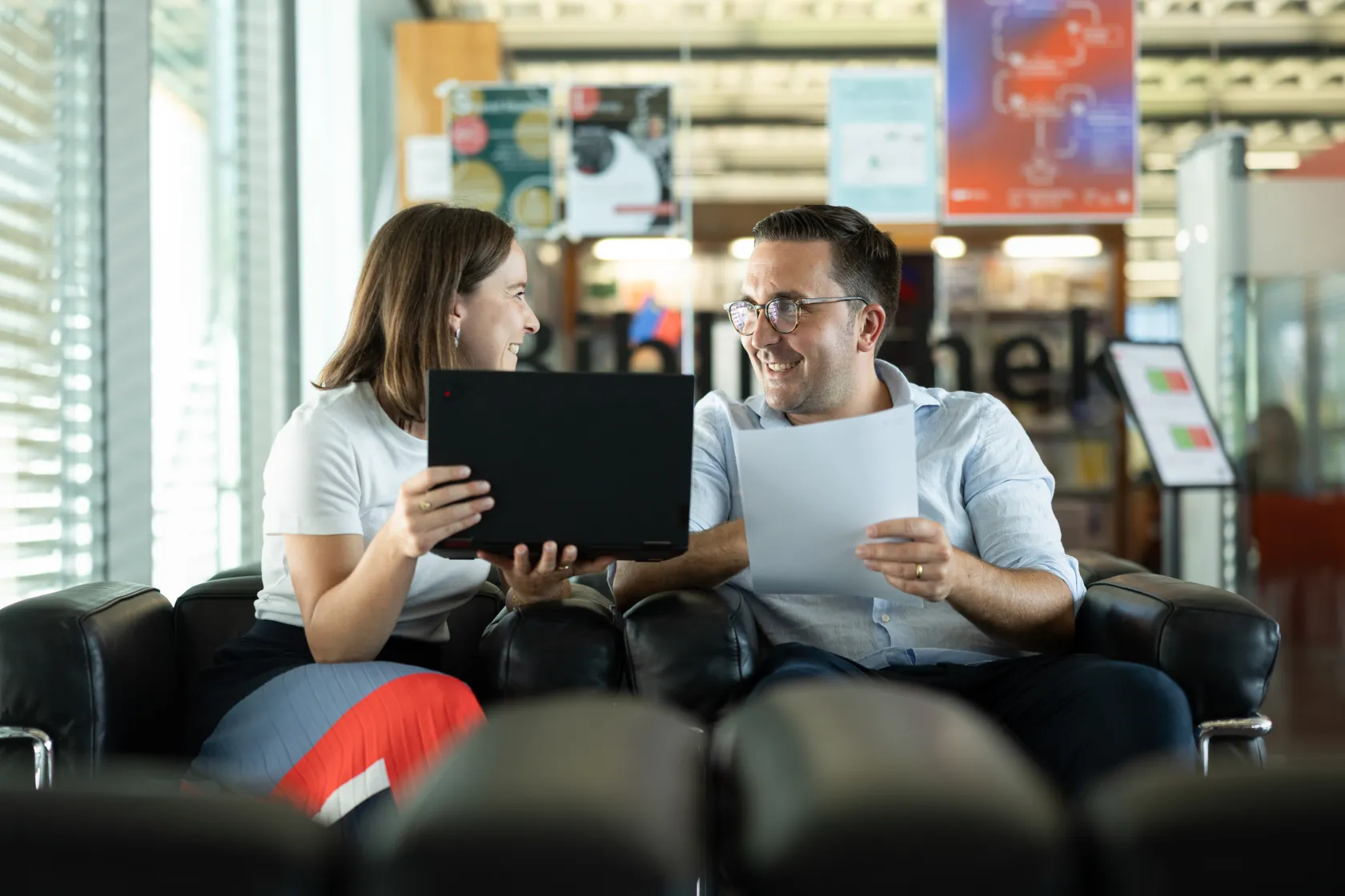 Zwei Personen lachen während eines Arbeitsgesprächs mit Laptop und Papierdokumenten im Lesebereich der Bibliothek.