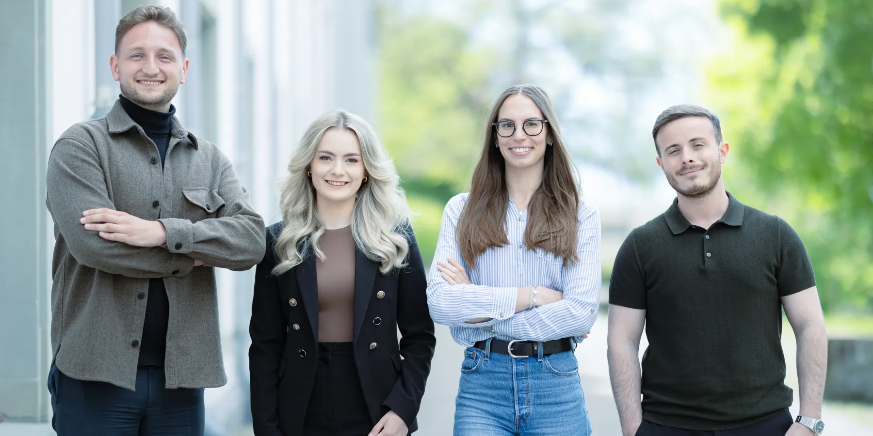Vier Studierende im Masterstudiengang Information Systems der Universität Liechtenstein stehen nebeneinander auf einem Gehweg vor einem Universitätsgebäude und lächeln in die Kamera. Die Kleidung reicht von Business- bis Casual-Stil.
