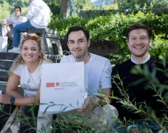 Drei neue Studierende sitzen auf dem Vorplatz des Campus Universität Liechtenstein und haben eine Tasche mit dem Logo der Universität in der Hand