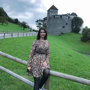 Studentin der Universität Liechtenstein sitzt vor dem Schloss Vaduz auf einem Holzzaun mit Blick in die Kamera, umgeben von grüner Hügellandschaft.