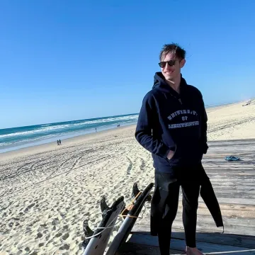 Student der Universität Liechtenstein steht barfuß am Strand mit Surfausrüstung und trägt einen Hoodie mit Universitätslogo während des Auslandssemesters.