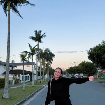 Studentin der Universität Liechtenstein steht lachend auf einer Straße mit Palmen in Australien und zeigt auf ihre Umgebung beim Auslandssemester.
