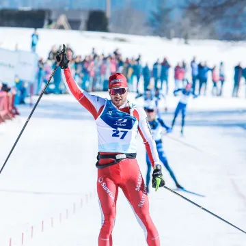 Ein Langläufer der Universität Liechtenstein überquert im roten Wettkampfanzug die Ziellinie eines Langlaufrennens im Schnee, jubelnd mit erhobenem Arm.