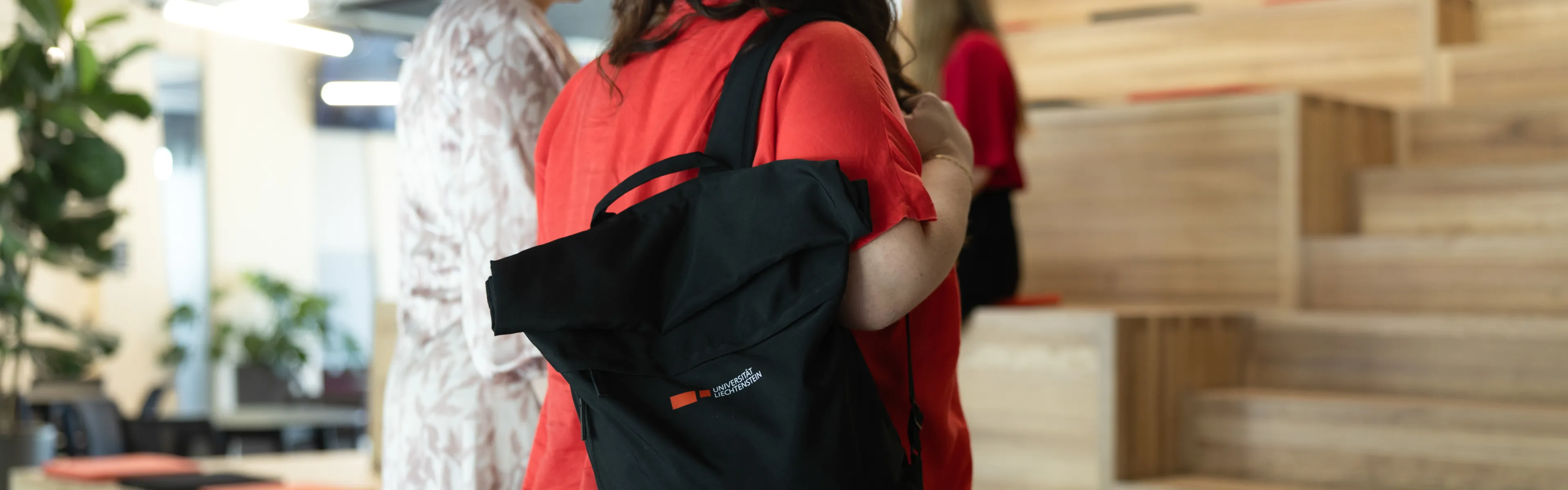 Studierende mit Uni-Rucksack stehen vor den Sitzstufen, im Hintergrund entspannte Gesprächsszene.