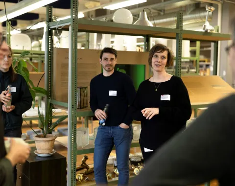 Eine Gruppe von Menschen steht in einem Innenraum vor Regalen mit Baumaterialien. Zwei Personen tragen Namensschilder und lächeln in die Kamera. Im Hintergrund sind Pflanzen und Lampen zu sehen.