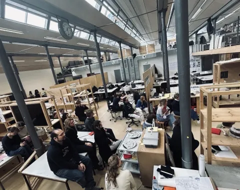 Ein großer, heller Raum mit hohen Decken in der Liechtenstein School of Architecture. Studierende sitzen an Tischen und hören einer Präsentation zu. Im Vordergrund sind Architekturmodelle und Zeichnungen zu sehen. Die Atmosphäre wirkt konzentriert und kreativ.