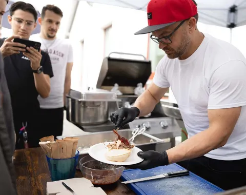 Ein Mann mit roter Kappe und schwarzen Handschuhen bereitet Essen auf einem Teller vor. Im Hintergrund stehen mehrere Personen und beobachten ihn. Eine Grillstation ist ebenfalls zu sehen. Die Szene findet im Freien unter einem Zelt statt.
