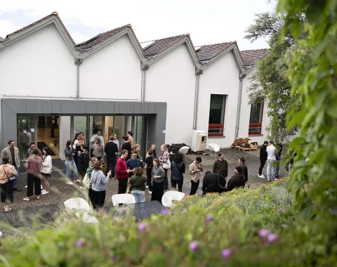 Eine Gruppe von Menschen versammelt sich im Innenhof der Liechtenstein School of Architecture während des Architektur Sommerfests. Im Hintergrund sind moderne Gebäude mit großen Fenstern zu sehen. Die Atmosphäre ist lebhaft und gesellig.