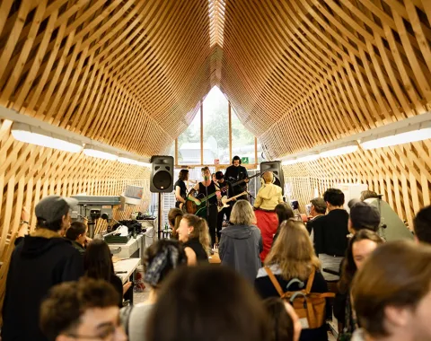Eine Band spielt vor einem Publikum in einem architektonisch beeindruckenden Raum mit Holzgitterstruktur bei einem Event der Universität Liechtenstein.