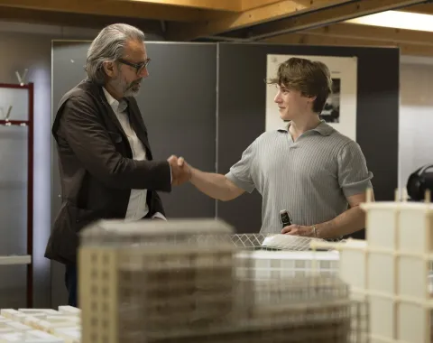 Zwei Personen schütteln sich die Hand in einem Raum mit Architekturmodellen. Eine Person hält ein Getränk. Die Szene vermittelt eine freundliche und professionelle Atmosphäre.