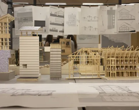 Architekturmodelle aus Holz und Papier stehen auf einem Tisch, umgeben von aufgehängten Architekturzeichnungen. Im Hintergrund sind unscharf Personen zu erkennen.