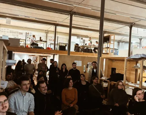 Eine Gruppe von Studierenden und Alumni sitzt in einem Architekturstudio und hört einer Präsentation zu. Im Hintergrund sind Arbeitsplätze und eine Uhr an der Wand zu sehen. Die Atmosphäre ist konzentriert und interessiert.