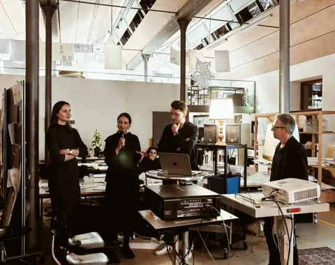 Eine Gruppe von Personen steht in einem modernen Architekturstudio. Eine Person spricht in ein Mikrofon, während andere aufmerksam zuhören. Im Raum sind technische Geräte und Präsentationsmaterialien zu sehen.