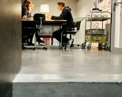 Drei Personen sitzen an einem Tisch in einem modernen Seminarraum und diskutieren. Im Hintergrund sind Architekturpläne an der Wand sichtbar. Der Raum ist hell beleuchtet und wirkt einladend.