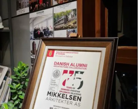 Ein gerahmtes Zertifikat zur Anerkennung der Zusammenarbeit im Bereich Architektur zwischen Dänemark und Pakistan, ausgestellt an Fasih D. Khan, steht auf einem Regal. Im Hintergrund sind Architekturprojekte und Gruppenfotos zu sehen.