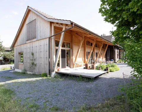 Ein modernes Holzgebäude mit schrägen Stützen und einer Terrasse, umgeben von Kies und Grünflächen, im ländlichen St. Galler Rheintal.