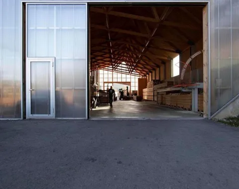 Ein modernes Gebäude mit einer großen offenen Tür, die den Blick auf eine helle, hölzerne Innenstruktur freigibt. Im Inneren sind Personen und gestapelte Holzmaterialien sichtbar. Die Architektur betont klare Linien und natürliche Materialien.
