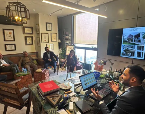 Ein Büro mit mehreren Personen, die in einem Meeting sitzen. Eine Person sitzt an einem Schreibtisch mit einem Laptop, während andere auf Stühlen und einem Sofa Platz genommen haben. An der Wand hängen Zertifikate und ein Bildschirm zeigt architektonische Entwürfe.