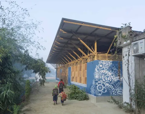 Ein modernes Gebäude mit einem Dach aus Bambus und Wellblech, verziert mit blauen, traditionellen Mustern. Im Vordergrund gehen mehrere Personen auf einem sandigen Weg entlang, umgeben von grüner Vegetation.