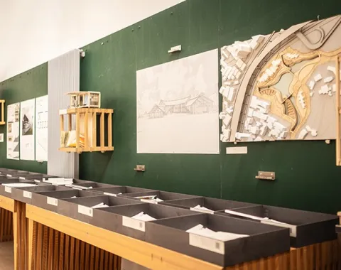 Architekturmodelle und Zeichnungen an einer grünen Pinnwand in einem Raum der Liechtenstein School of Architecture. Im Vordergrund sind Ablagefächer mit Dokumenten sichtbar.