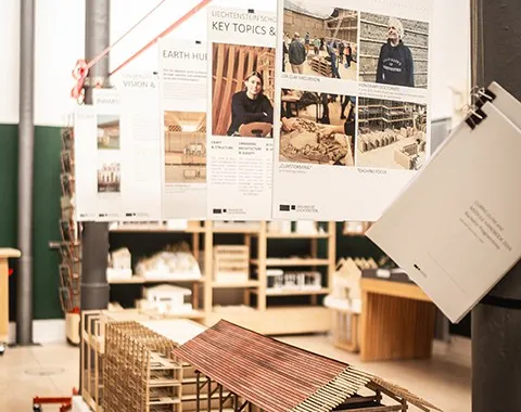 Ein Poster der Liechtenstein School of Architecture zeigt die "Key Topics & Units" mit einem Bild von zwei Personen vor architektonischen Modellen. Im Hintergrund sind weitere Poster zu sehen, die verschiedene Architekturthemen präsentieren.