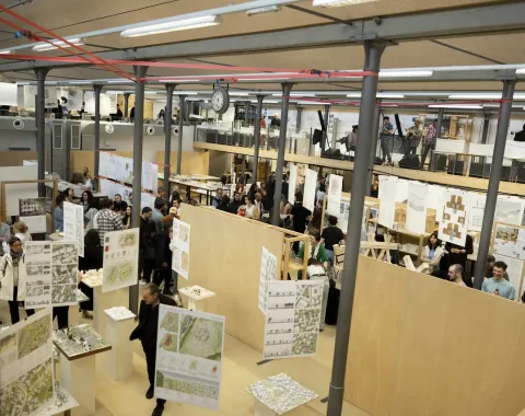 Eine große, gut besuchte Ausstellungshalle mit zahlreichen Architekturmodellen und Plänen, die an Holzwänden präsentiert werden. Menschen bewegen sich durch den Raum, betrachten die ausgestellten Werke und diskutieren. Die Atmosphäre ist lebhaft und interessiert.