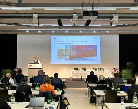 Ein Vortragender steht an einem Rednerpult vor einer Leinwand, auf der eine Präsentation gezeigt wird. Im Publikum sitzen mehrere Personen an Tischen mit Laptops. Die Veranstaltung findet in einem modernen Konferenzraum statt.