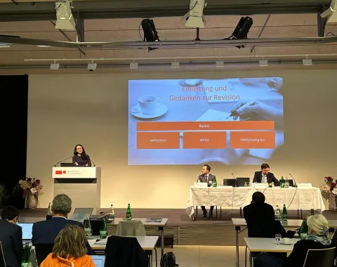 Eine Person hält einen Vortrag auf einer Bühne beim Liechtensteinischen Bankrechtsforum an der Universität Liechtenstein. Im Hintergrund ist eine Präsentationsfolie mit dem Titel "Einleitung und Gedanken zur Revision" zu sehen. Zwei weitere Personen sitzen an einem Tisch auf der Bühne. Im Publikum sind mehrere Zuhörer.