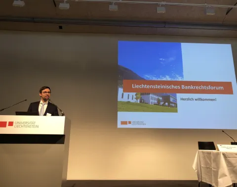 Ein Redner steht am Podium der Universität Liechtenstein während des Liechtensteinischen Bankrechtsforums. Auf der Leinwand ist eine Präsentationsfolie mit der Aufschrift „Liechtensteinisches Bankrechtsforum – Herzlich willkommen!“ zu sehen.