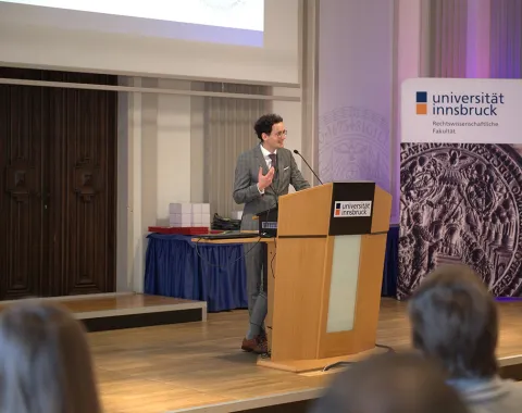Ein Mann hält eine Präsentation an einem Rednerpult in einem Saal der Universität Innsbruck. Im Hintergrund sind ein Banner der Universität und ein Tisch mit Gegenständen zu sehen. Publikum sitzt im Vordergrund.