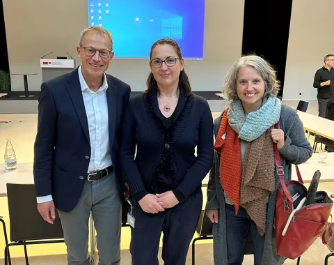 Drei Personen stehen lächelnd in einem Auditorium der Universität Liechtenstein. Im Hintergrund sind ein Rednerpult und eine Leinwand mit einer Präsentation zu sehen.