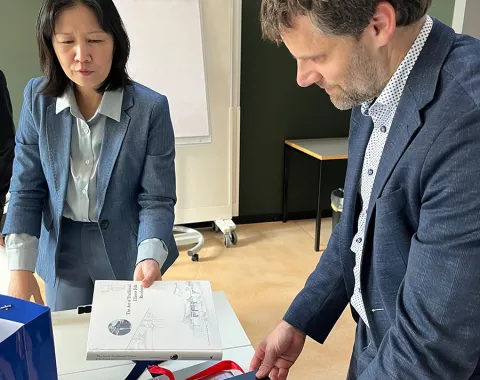 Zwei Personen in formeller Kleidung betrachten Bücher auf einem Tisch in einem Konferenzraum. Im Hintergrund ist ein Flipchart zu sehen.