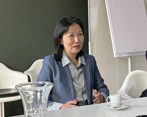 Eine Person in einem blauen Blazer sitzt an einem Konferenztisch mit einer Tasse Kaffee und einem Notizblock. Im Hintergrund ist ein Flipchart zu sehen.